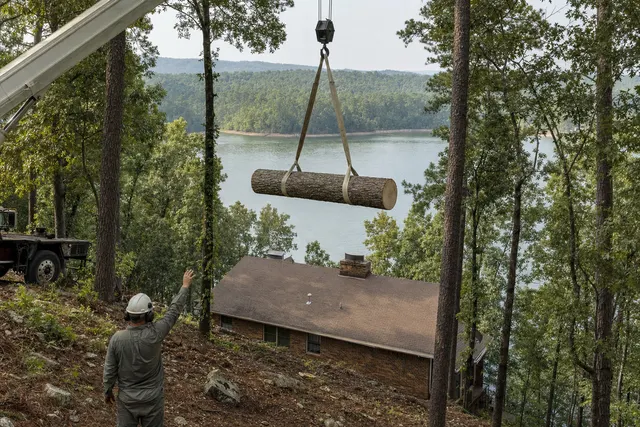 Pine trunk section lifted level by a crane over a lake house on a steep wooded lot